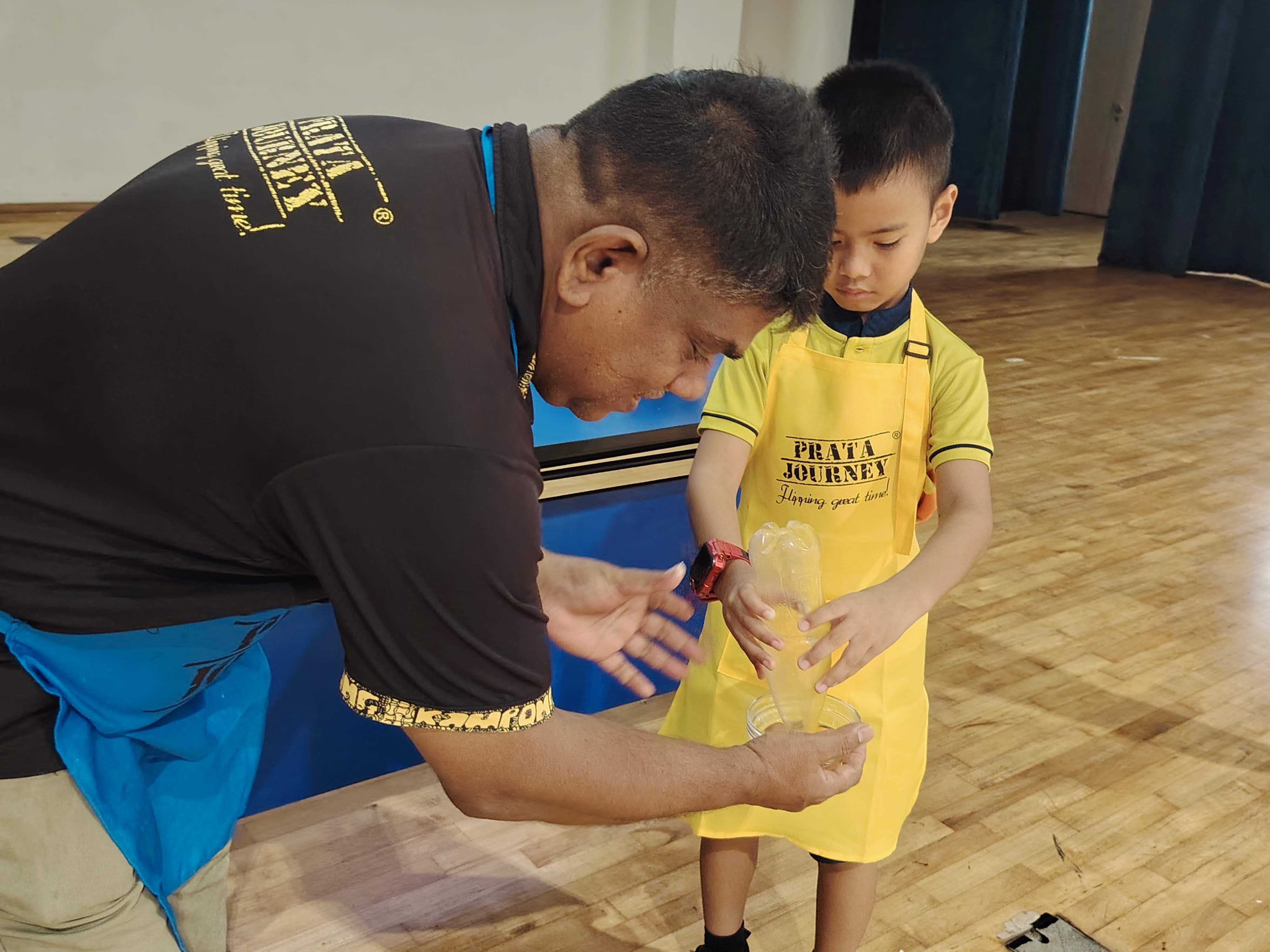 A roti prata instructor demonstrating to a student on how to suck up an egg using a plastic bottle